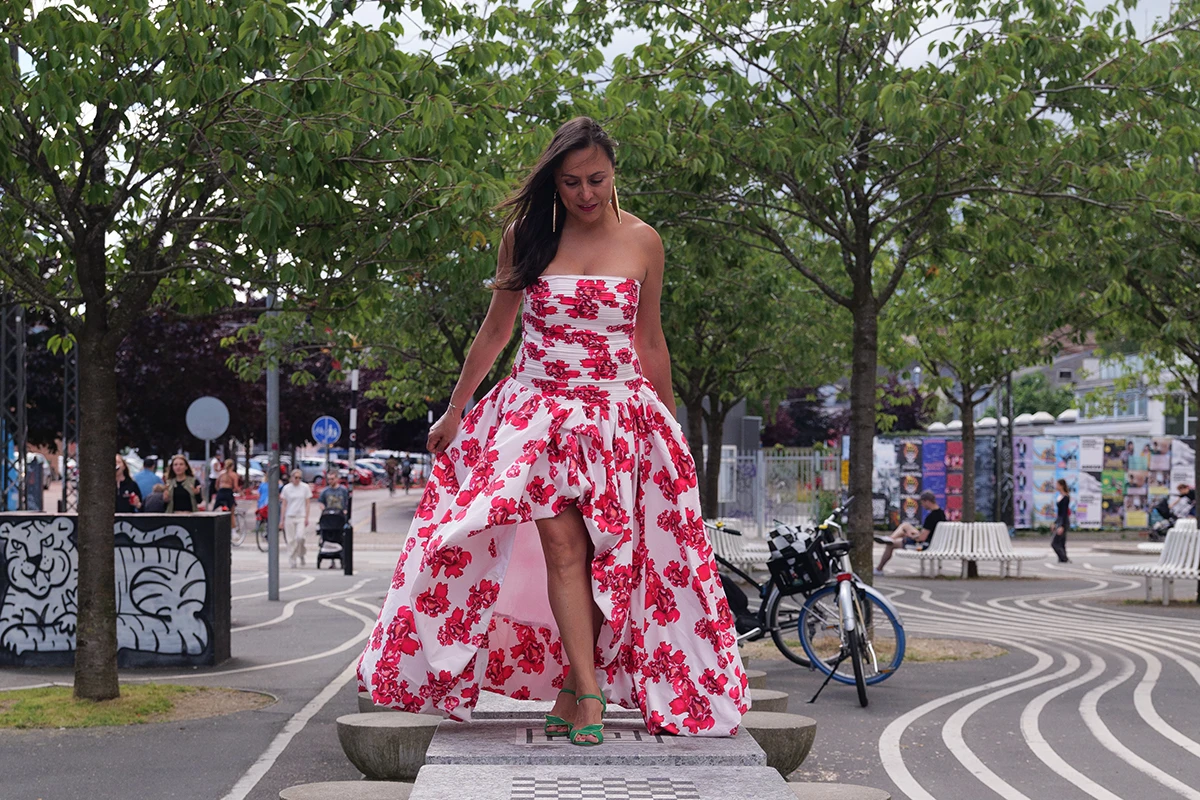 Full shot of latin woman in fashionable dress walking using the chess boards of the park as her catwalk. Surrounded by green summer cherry trees.
