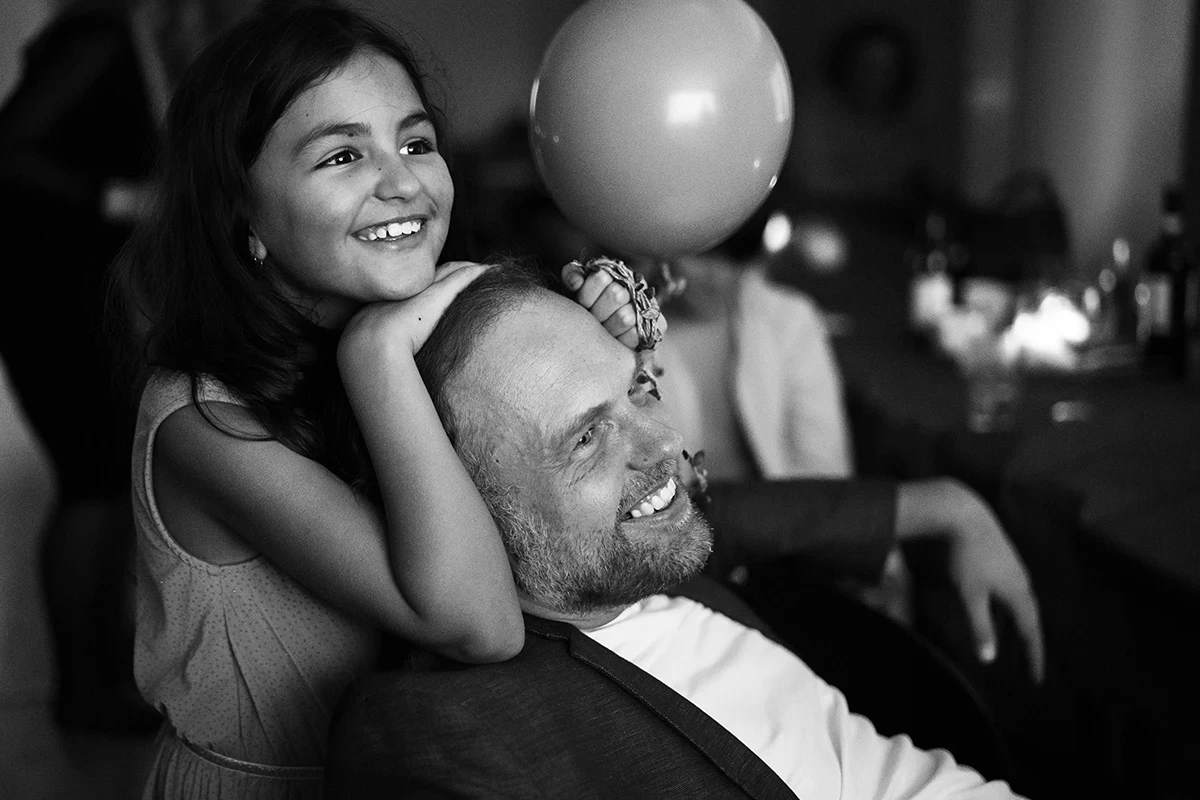 Father and daughter enjoying party entertainment; she stands behind him, chin resting on his head, both smiling warmly as they look ahead.